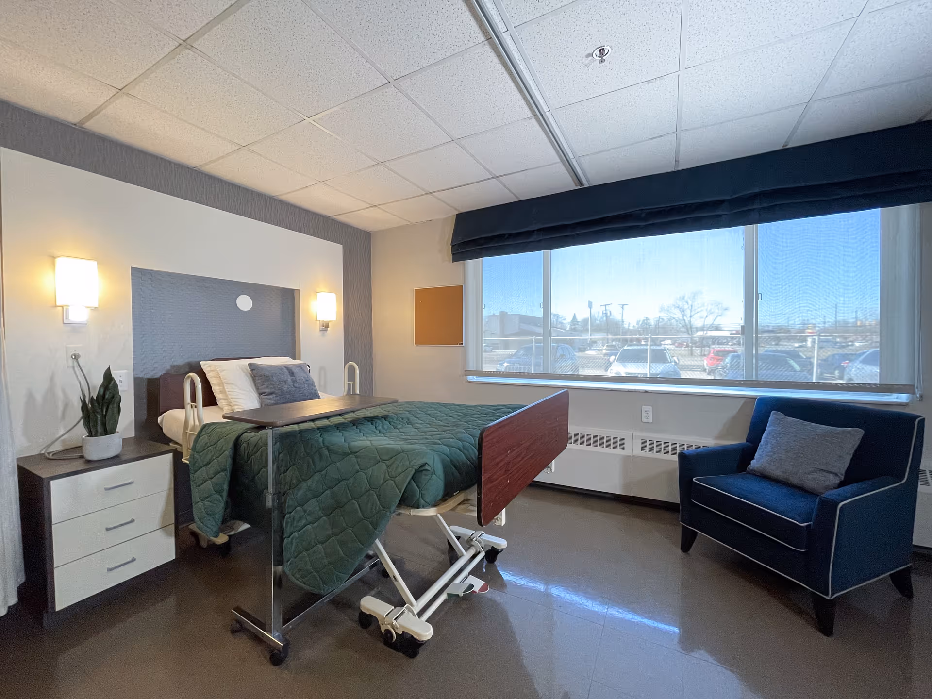 A bright resident bedroom with an adjustable hospital-style bed, overbed table, bedside drawers, an armchair and a large window.
