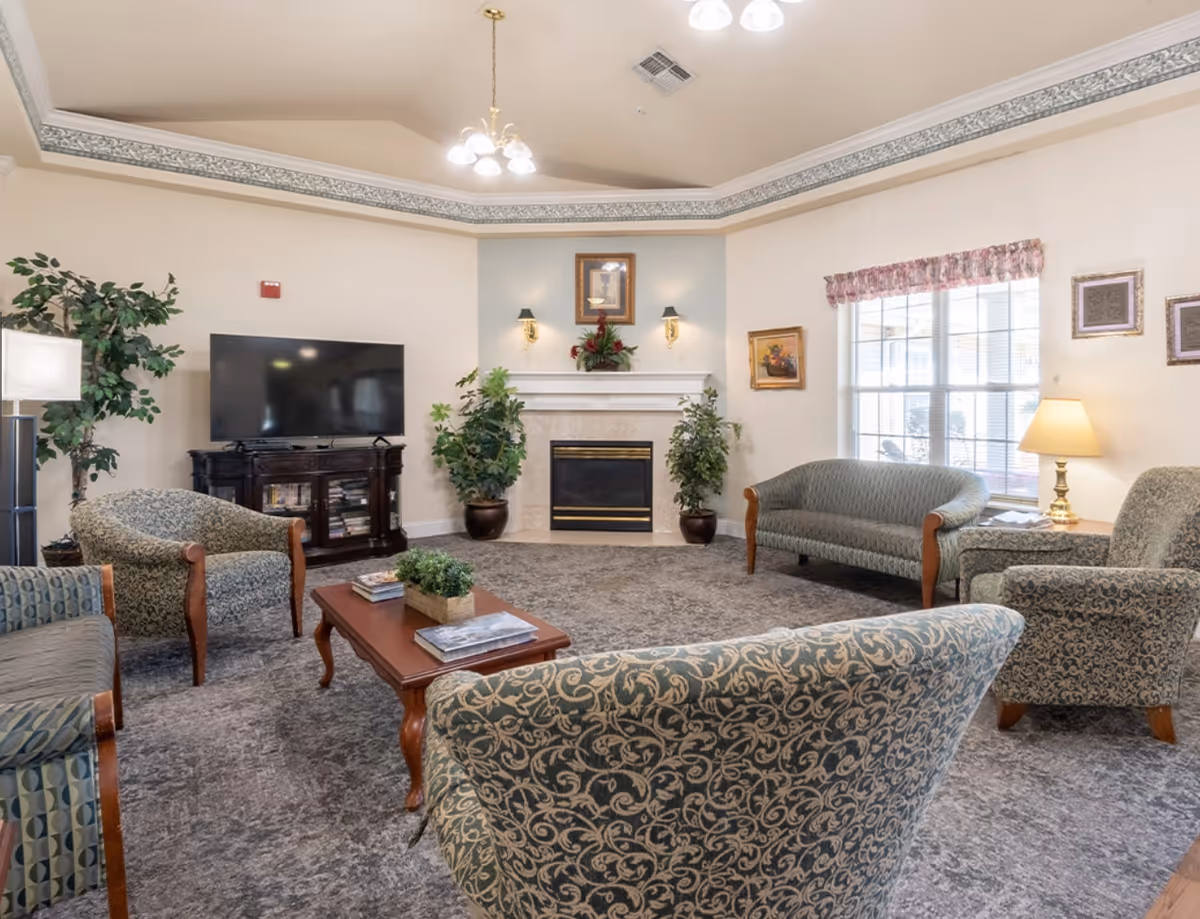 Spacious common living room with upholstered chairs and sofas arranged around a coffee table, a fireplace, TV and large window.