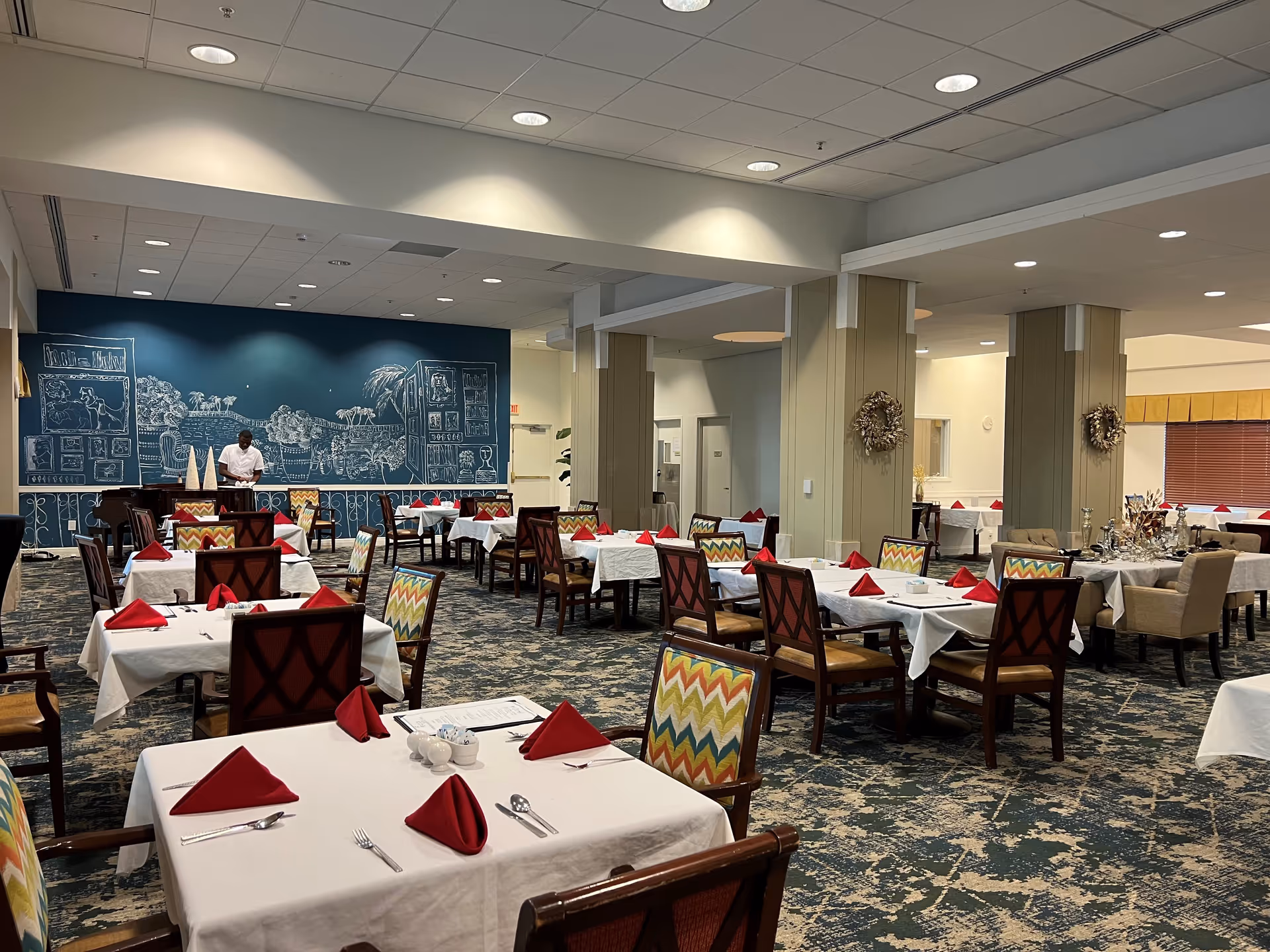 A spacious dining room with multiple tables covered in white tablecloths and set with red folded napkins, silverware, and small condiment containers. The chairs have colorful zigzag patterned upholstery. A person is standing near the back wall, which features a large blue mural with white chalk-like drawings of various scenes and objects. The room has a patterned carpet and neutral-colored walls with columns decorated with wreaths.