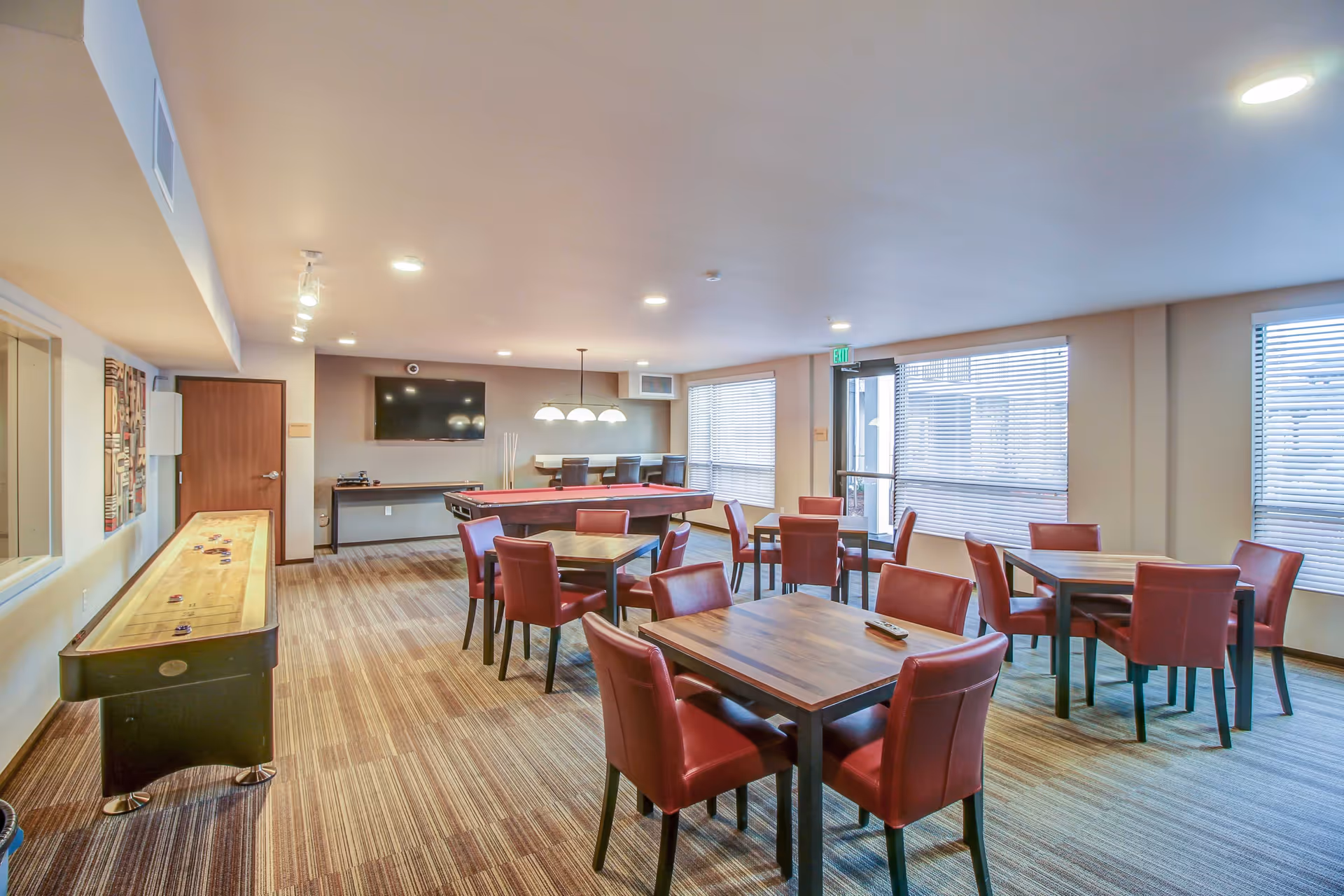 A spacious recreational room with several wooden tables and red cushioned chairs arranged neatly. The room features a shuffleboard table on the left and a pool table in the center back. A large flat-screen TV is mounted on the far wall above a long console table. Large windows with blinds allow natural light to fill the room, and there is a door with an exit sign above it on the right side.