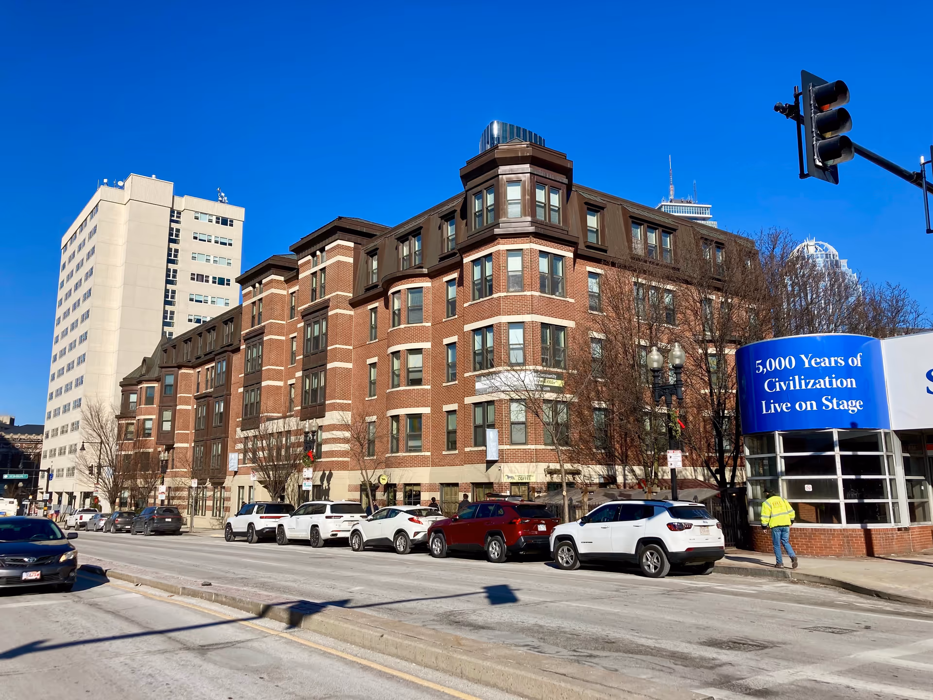 Street view of a multi-story red brick residential building with parked cars along the curb and a blue promotional sign at the corner.