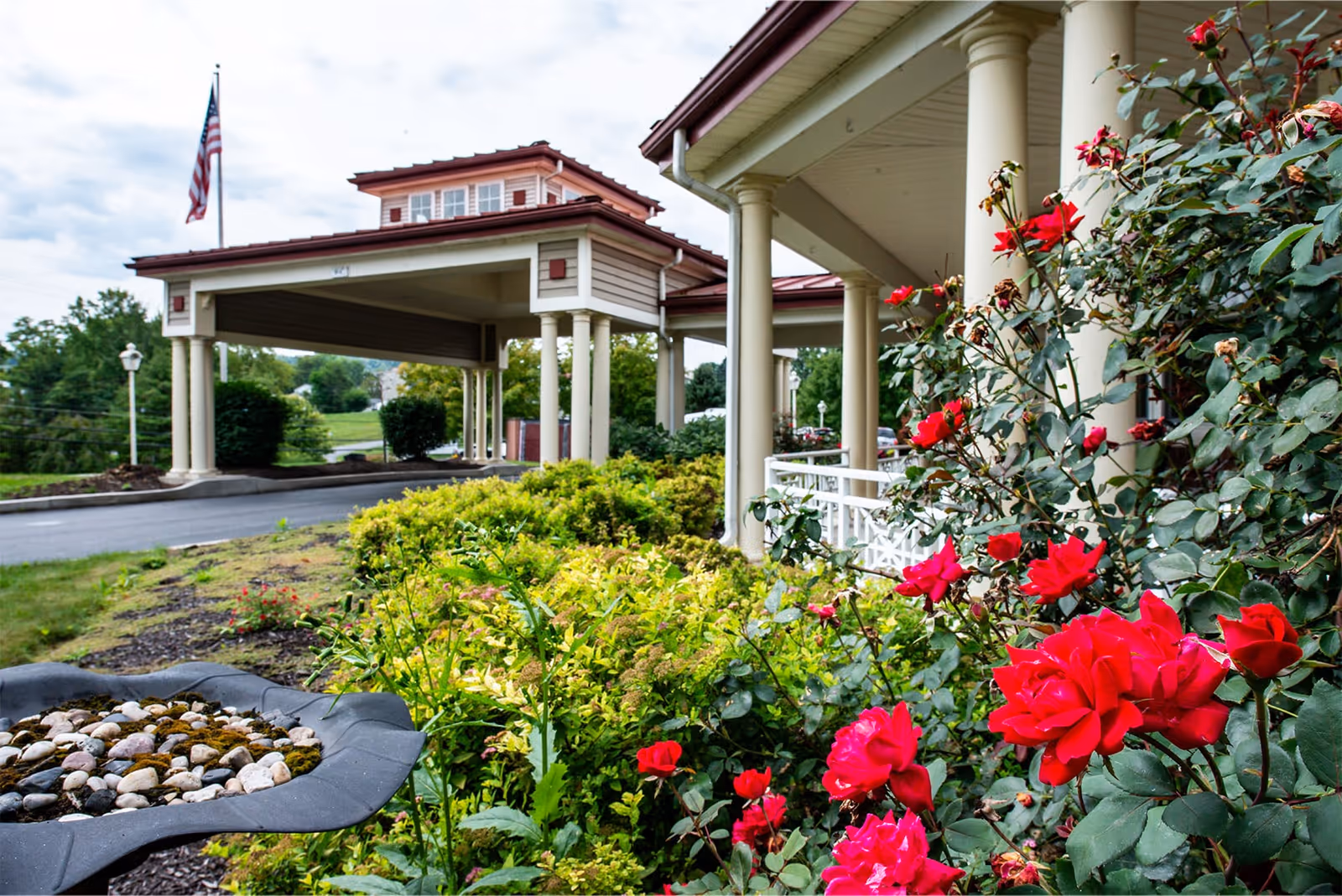 View of the entrance to The Winds At Mattern Orchard by New Perspective, featuring a covered driveway with columns, an American flag, and a garden with vibrant red roses and green shrubs in the foreground.