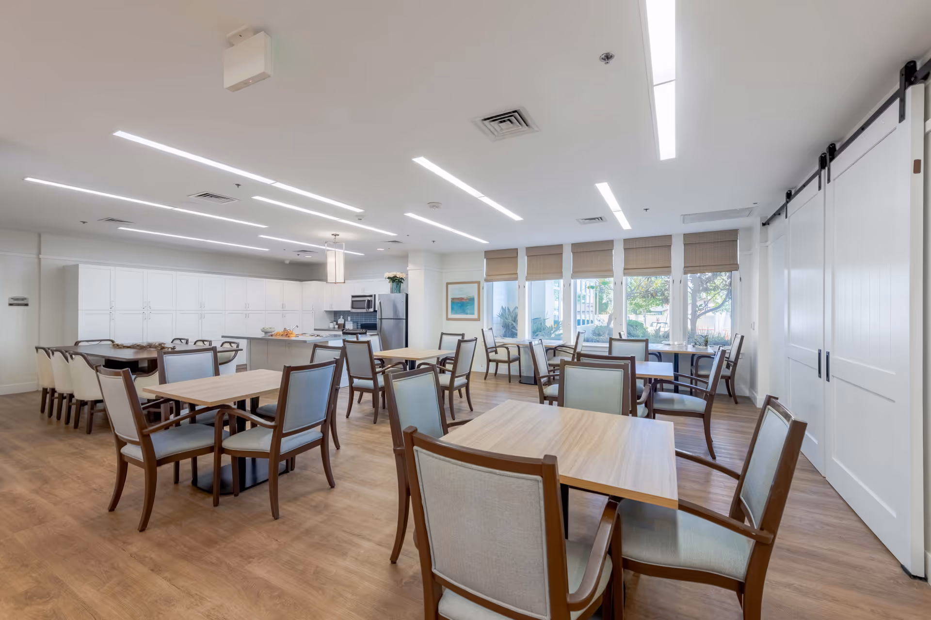 A bright and spacious dining room with multiple wooden tables and cushioned chairs arranged neatly. The room features large windows with beige blinds allowing natural light to fill the space. There is a modern kitchen area in the background with white cabinetry, a stainless steel refrigerator, microwave, and an island with a light fixture above it. The flooring is wood, and the ceiling has recessed linear lighting.