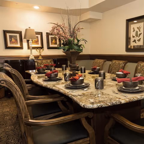 Elegant dining room with a large marble table set for six people with dark woven bowls, plates, silverware, and red cloth napkins. The room features upholstered chairs, a floral centerpiece in a tall vase, framed artwork on the walls, and a sideboard with a lamp and decorative items.