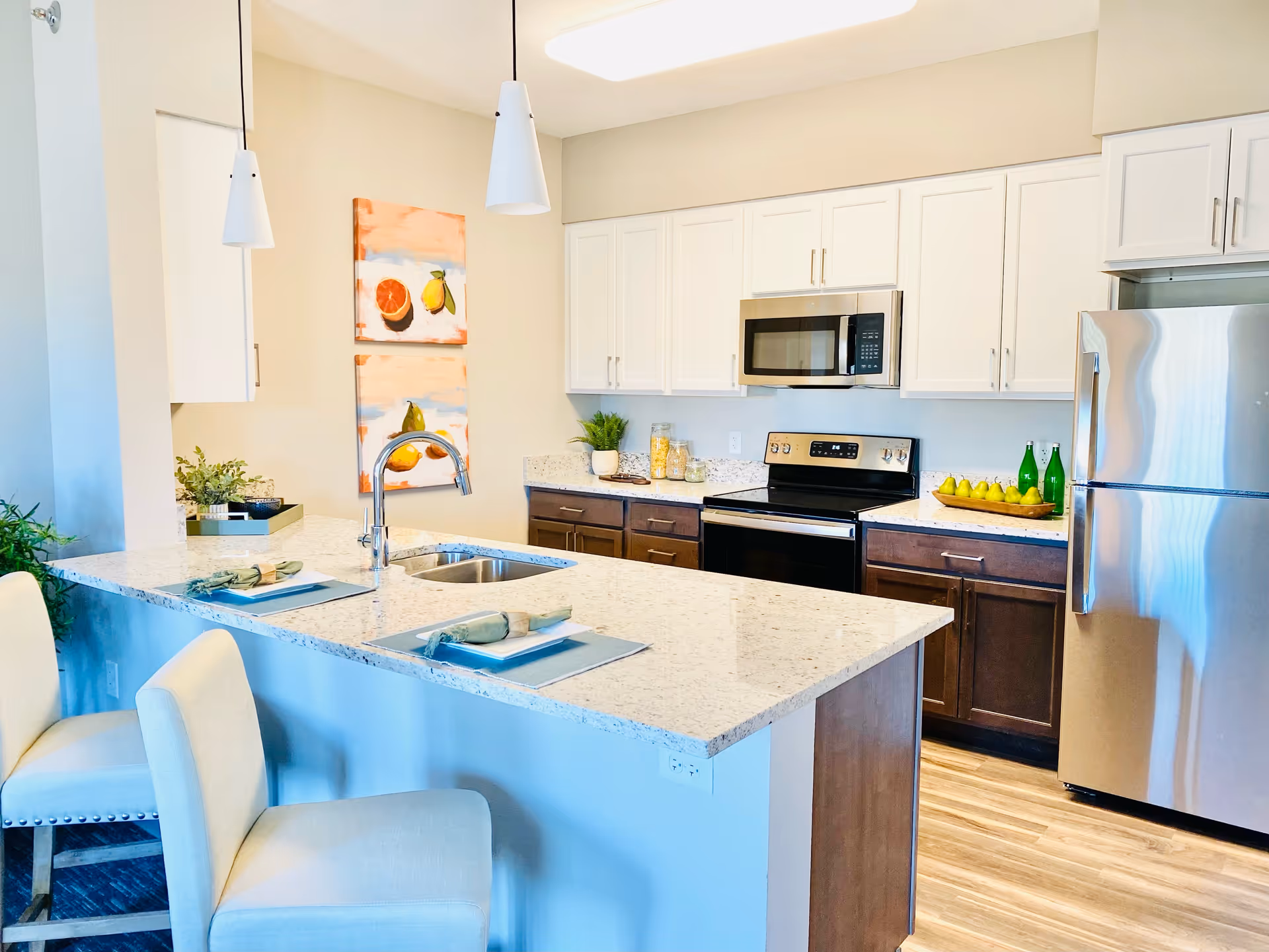 Modern kitchen with a large white granite island featuring a double sink and two place settings with napkins and plates. The kitchen has white upper cabinets, dark lower cabinets, stainless steel appliances including a refrigerator, stove, and microwave. Two pendant lights hang above the island, and there are decorative paintings and plants on the countertops.