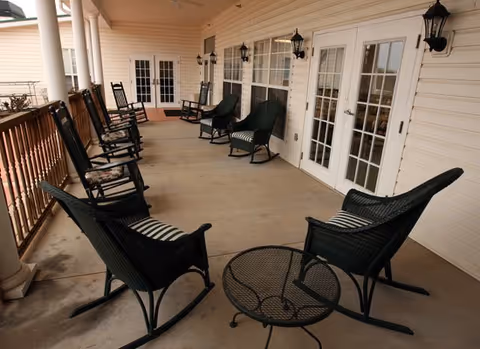 Covered outdoor porch area with multiple black rocking chairs and wicker chairs arranged along the wall and railing. There is a small round metal table in the foreground and several wall-mounted lantern-style lights. The porch has a beige exterior with white trim and multiple glass doors leading inside.