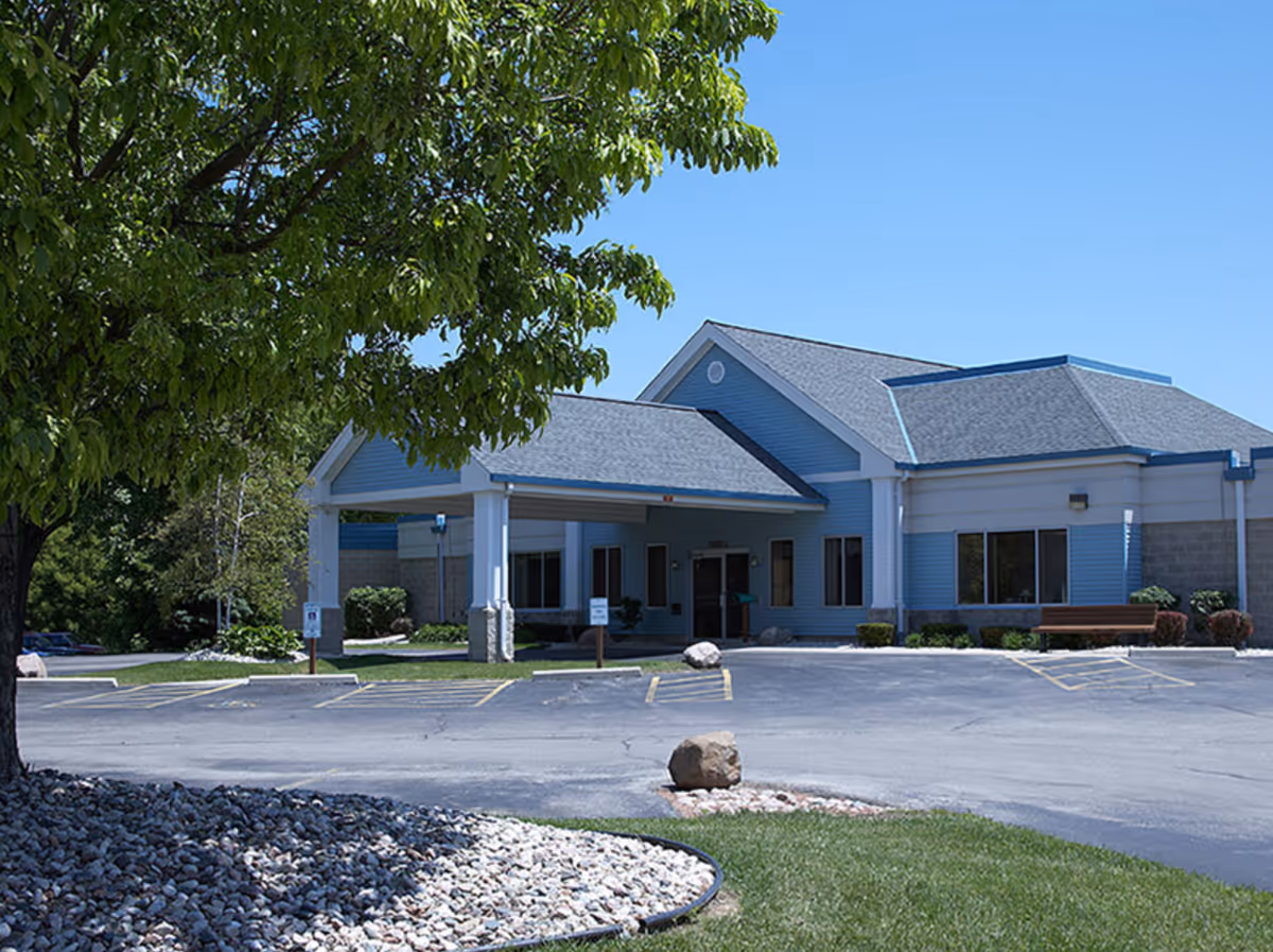 Exterior view of a single-story building with blue siding and a gray roof under a clear blue sky. The building has a covered entrance with white pillars and a parking lot in front. There is a large green tree on the left side and landscaped rocks and grass in the foreground.
