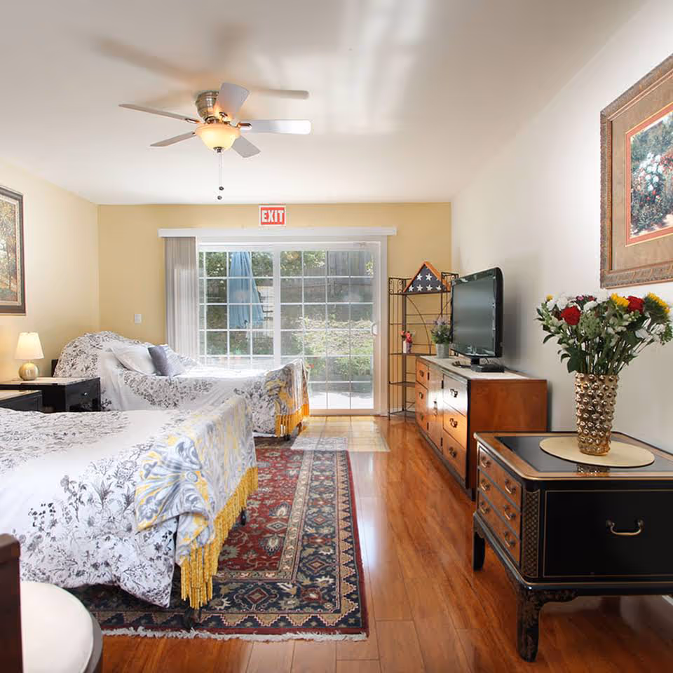 A bright bedroom with two beds covered in white and gray floral bedding, a ceiling fan, a patterned area rug, a wooden dresser with a flat-screen TV, a small black side table with a vase of colorful flowers, and a large sliding glass door leading outside.