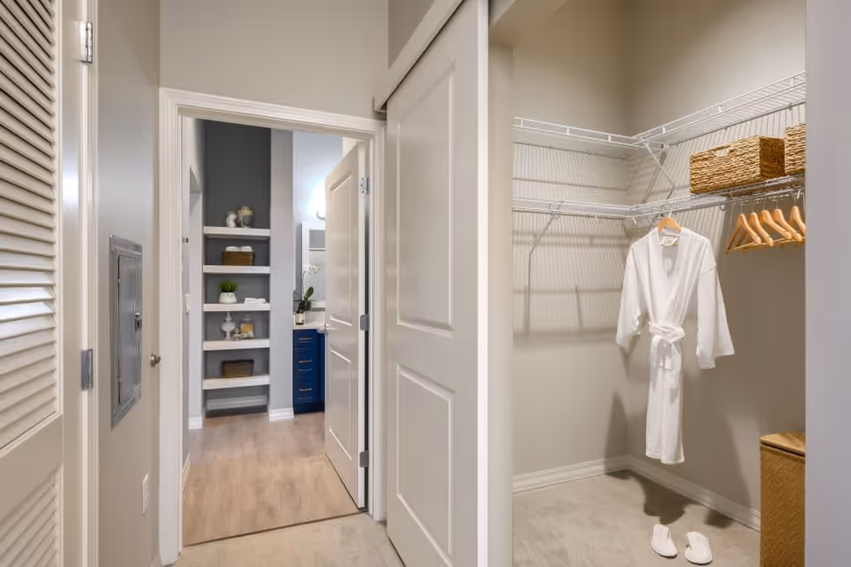 View of a walk-in closet with white wire shelving, a white robe hanging on a wooden hanger, and slippers on the floor. The closet is adjacent to a hallway leading to a bathroom with open shelving and a blue vanity cabinet.
