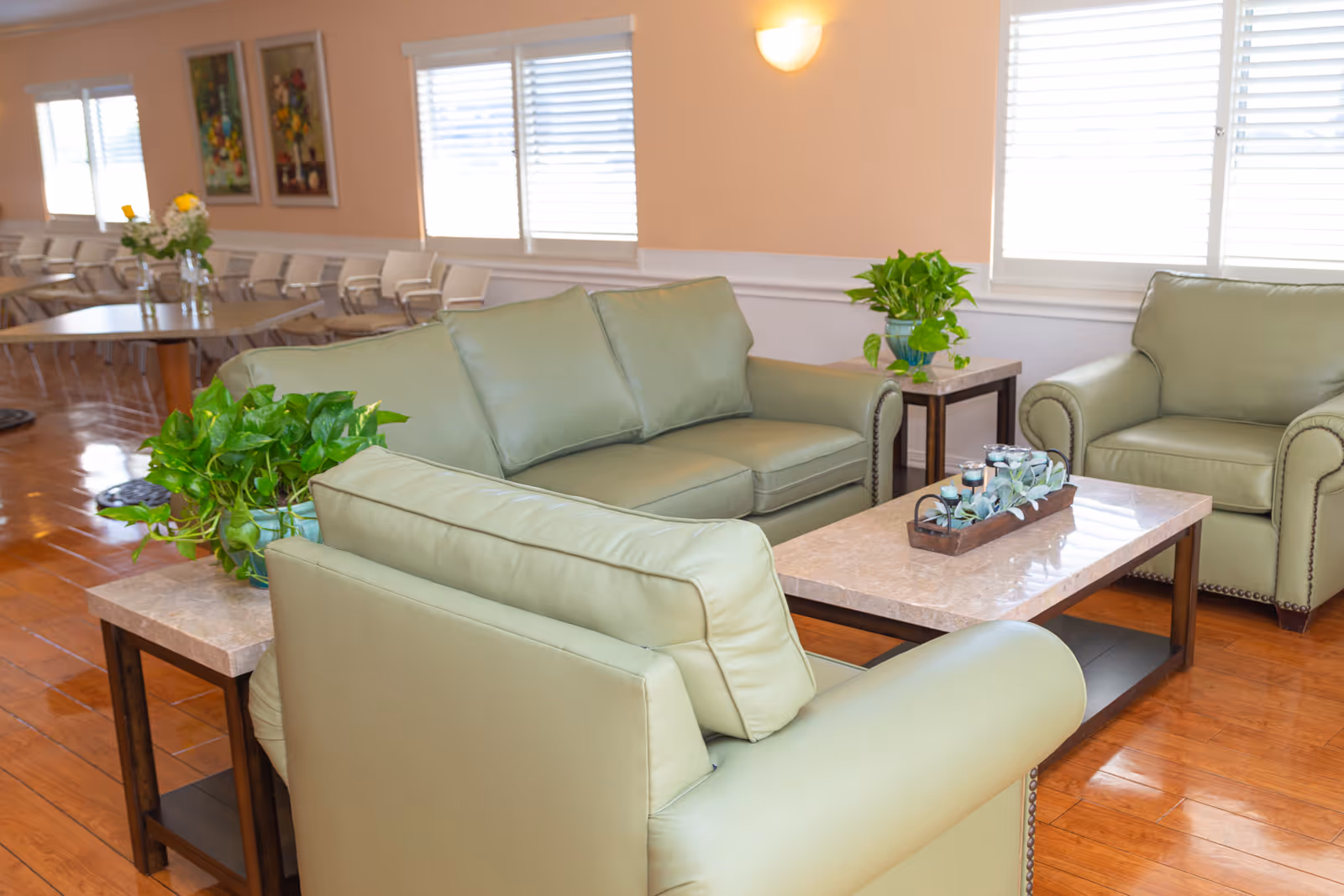 A bright and clean living room area with light green leather sofas and armchairs arranged around a rectangular marble coffee table. The coffee table has a decorative tray with candles and greenery. There are two side tables with green potted plants. The room has wooden flooring, large windows with white blinds, and soft peach-colored walls with framed artwork and wall sconces.