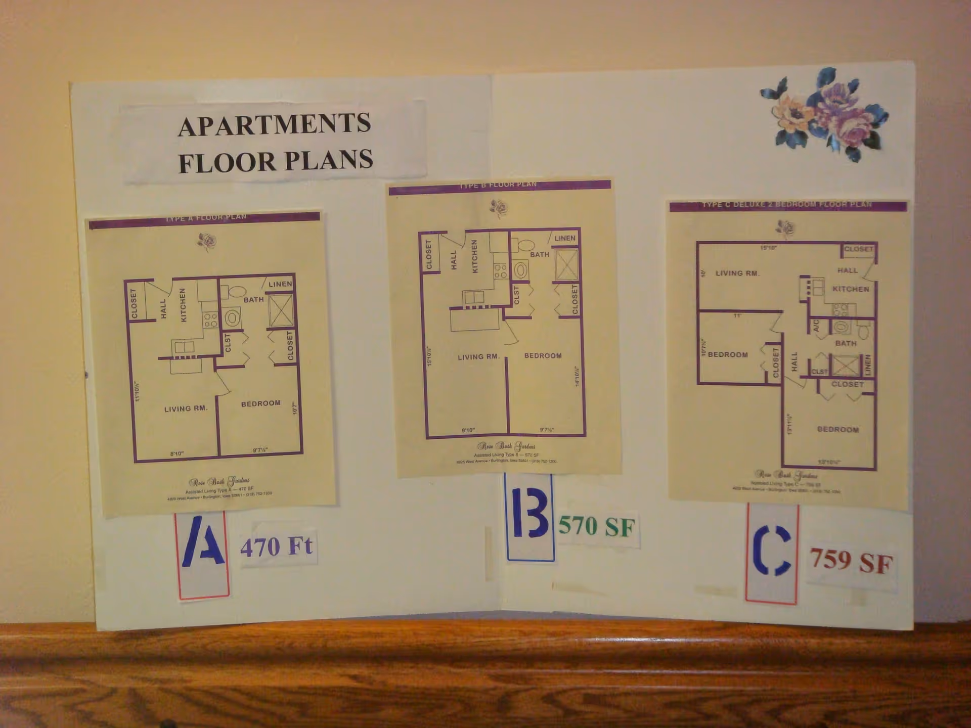 A display board titled "APARTMENTS FLOOR PLANS" showing three printed apartment floor plan diagrams labeled A 470 Ft, B 570 SF, and C 759 SF mounted above a wooden ledge.