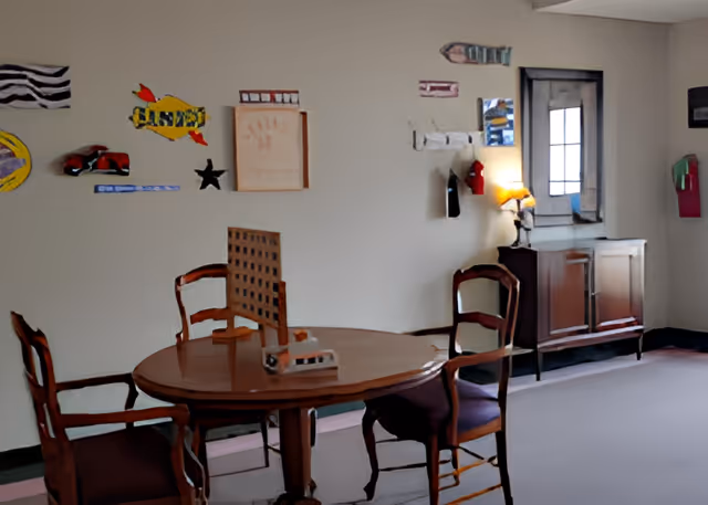 A small room with a round wooden table and four wooden chairs with purple cushions. On the table is a wooden pegboard game. The walls are decorated with colorful, abstract art pieces. There is a wooden sideboard with a lamp and a window with a reflective surface in the background.