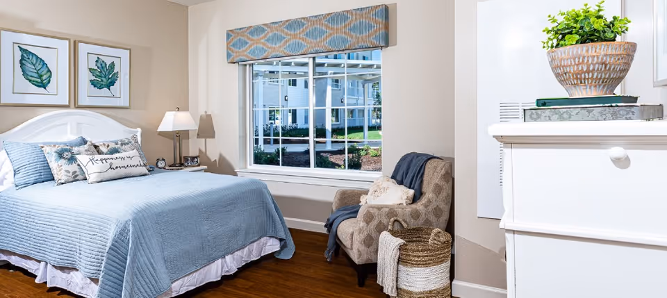 A cozy bedroom with a neatly made bed covered in a light blue quilt and multiple pillows, including one with the words 'Happily homemade'. Two framed leaf prints hang above the bed. Next to the bed is a nightstand with a lamp and an alarm clock. A window with a patterned valance shows an outdoor view of a building and greenery. An upholstered armchair with a throw blanket and pillow sits near a woven basket. A white dresser with a potted plant and books is partially visible on the right.