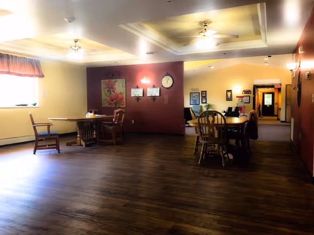 Interior view of a senior living facility common area with wooden floors, multiple wooden tables and chairs, a window with a valance, wall decorations including a large floral painting and a clock, and ceiling fans with lights.