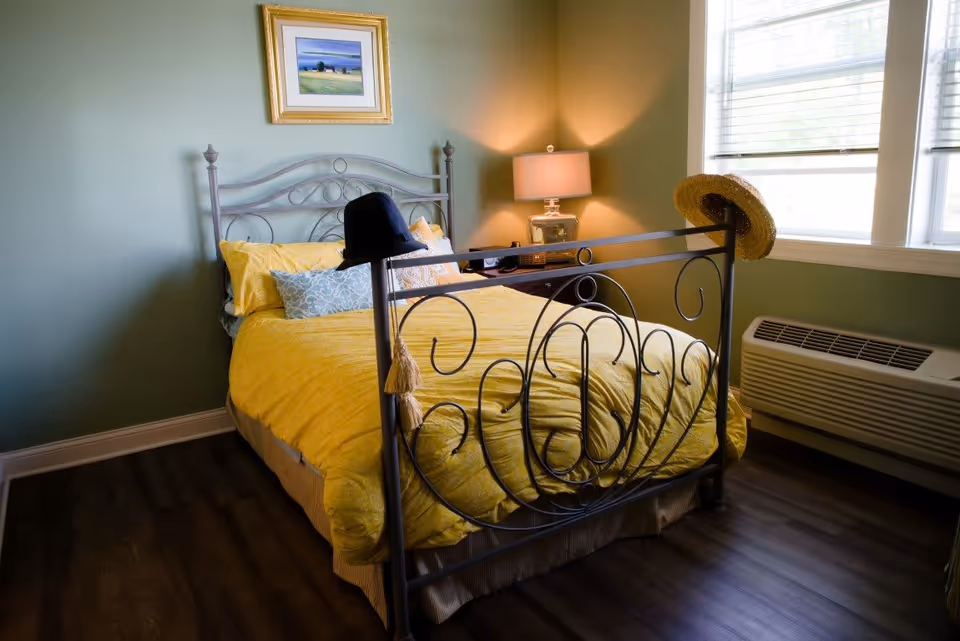 Sunlit bedroom with a wrought-iron bed dressed in yellow bedding, a bedside lamp, window, and wall art.