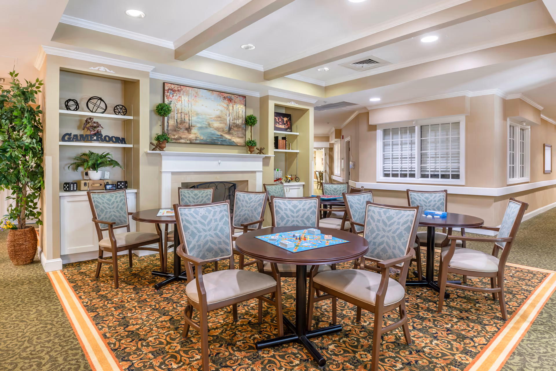 A cozy game room area in a senior living facility with several round tables and cushioned chairs arranged on a patterned carpet. Board games are set up on the tables. The room features built-in shelves with decorative items, a fireplace with a landscape painting above it, and potted plants. The walls are painted beige with white trim, and there is a window with white blinds on the right side.