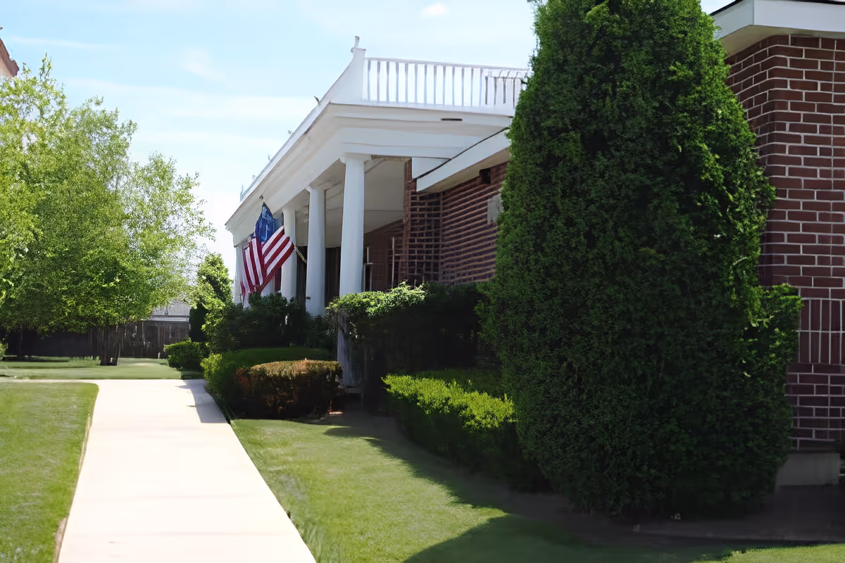 Side view of a brick building with white columns and an American flag hanging near the entrance. The building is surrounded by green bushes, trees, and a well-maintained lawn with a concrete walkway leading up to it.