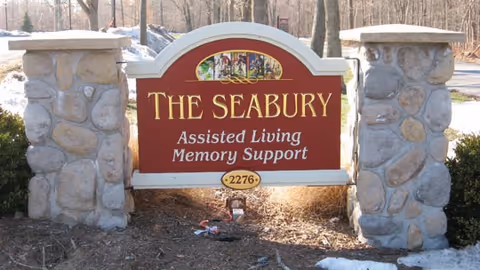 A stone and wood sign for The Seabury Assisted Living Memory Support, with the number 2276 displayed at the bottom. The sign is set outdoors with trees and a road in the background.