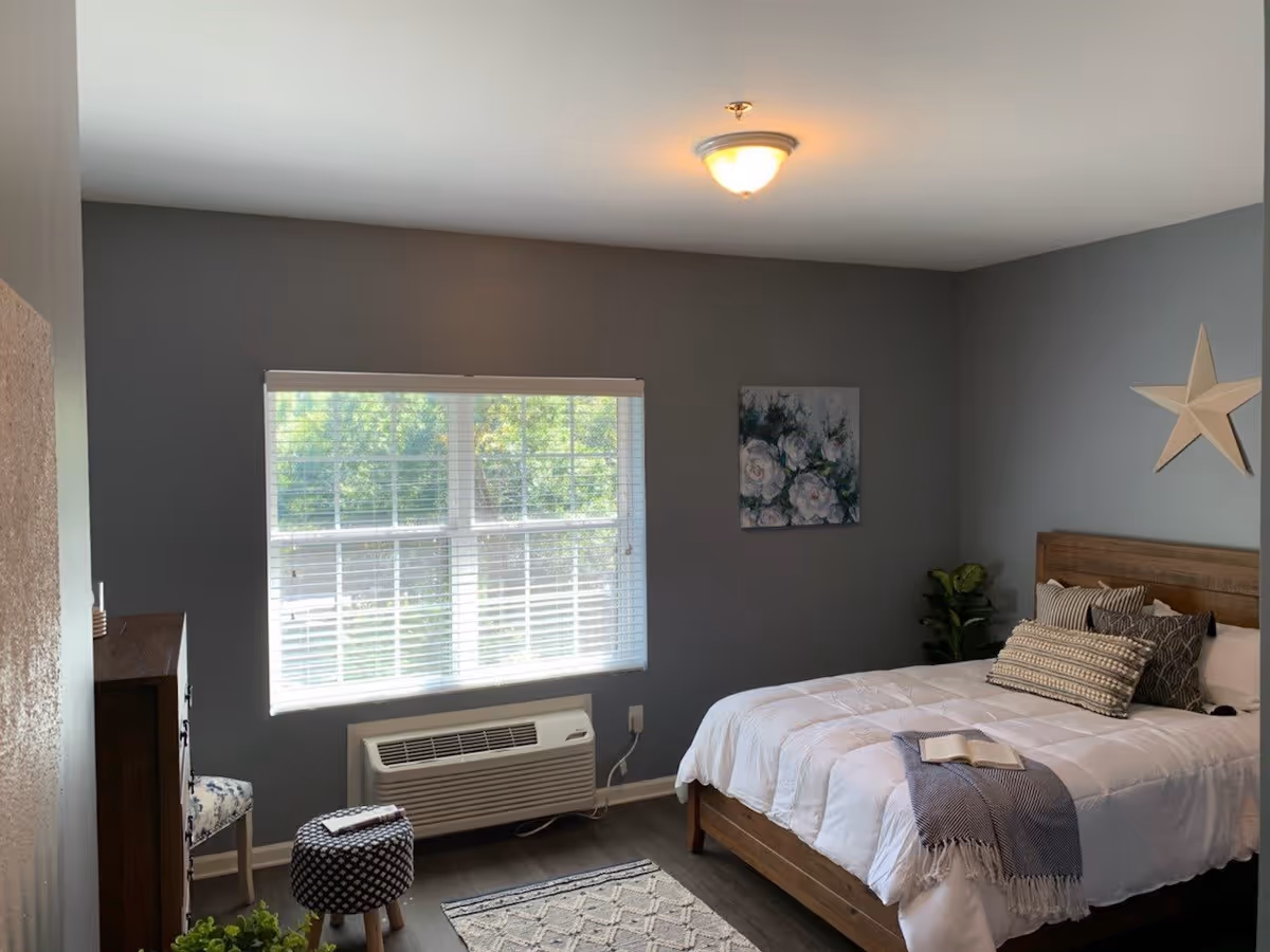 A cozy bedroom with gray walls, a large window with white blinds, a wooden bed with white bedding and multiple pillows, a small plant in the corner, a star decoration on the wall, a floral painting, a patterned ottoman, a small rug, and a wooden dresser partially visible.