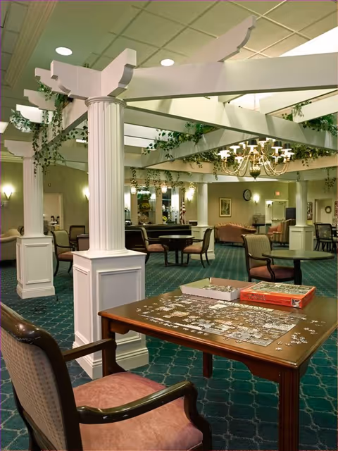 A spacious common area in a senior living facility featuring a green carpet with a diamond pattern, white decorative columns, and a white pergola structure with hanging greenery. Several tables and chairs are arranged throughout the room. In the foreground, a wooden table holds an incomplete jigsaw puzzle with puzzle pieces scattered around and the puzzle box nearby. The room is softly lit with wall sconces and a chandelier, and there are couches and additional seating in the background.