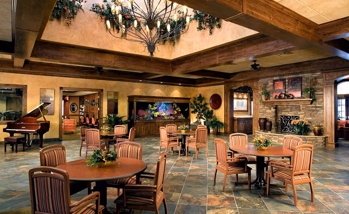 Spacious senior living common area with round wooden tables and striped cushioned chairs arranged on a tiled floor. The room features a large stone fireplace with decorative items on the mantel, a grand piano on the left side, and a large built-in aquarium with colorful fish. The ceiling has wooden beams and a decorative chandelier with greenery hanging from it. The walls are painted in warm tones with plants and framed artwork adding to the cozy ambiance.