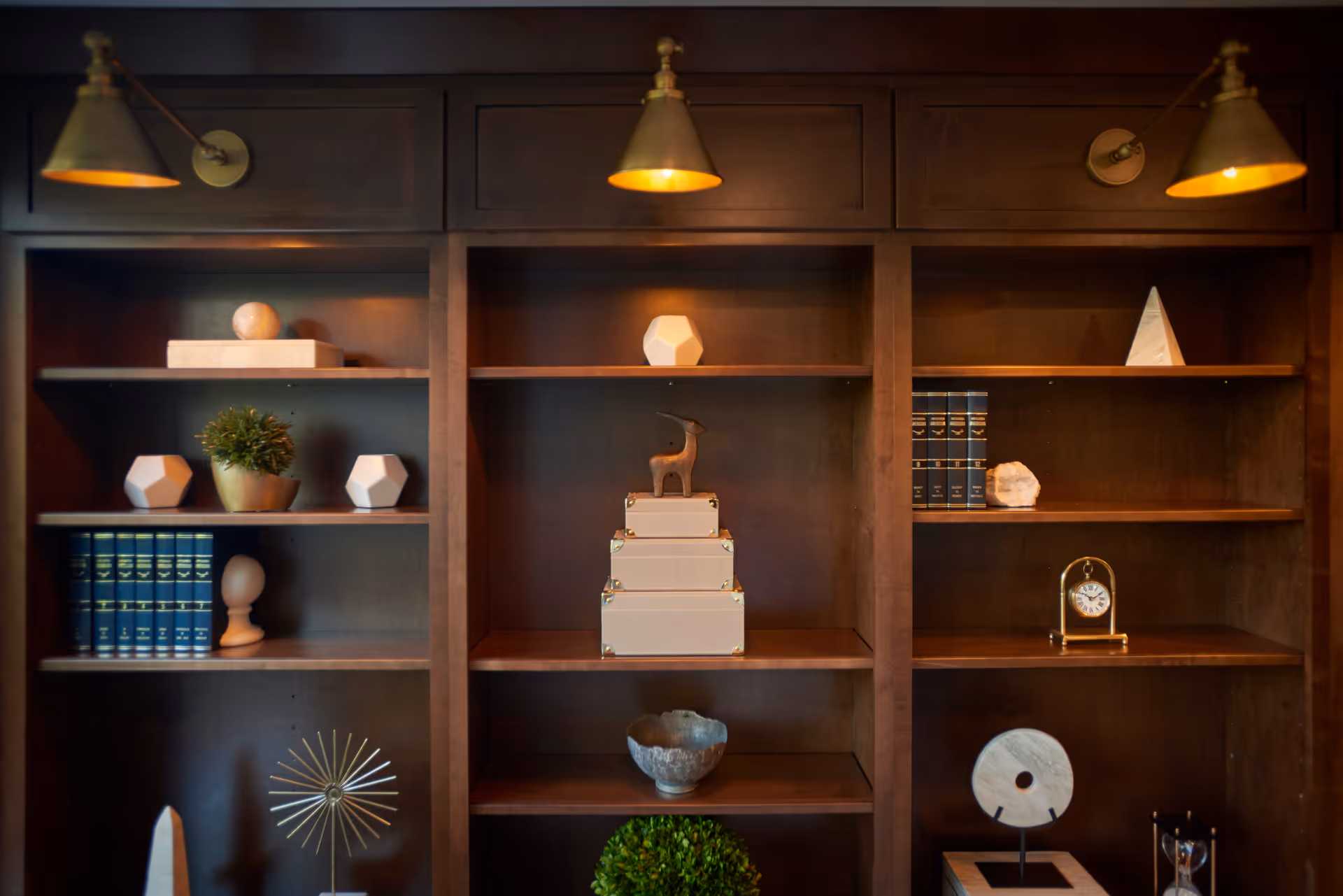 Wooden built-in shelving unit filled with decorative objects and topped by three brass wall lamps.