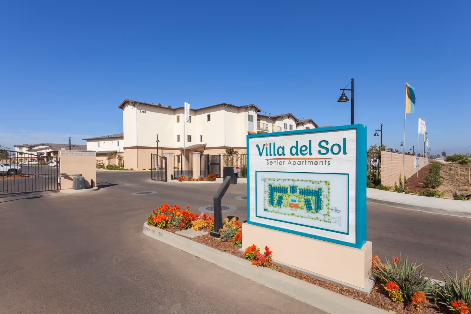 Entrance to Villa del Sol Senior Apartments with a large sign displaying the facility name and a site map. The gated driveway leads to a multi-story beige building under a clear blue sky. There are flower beds with red and yellow flowers near the sign and along the driveway.