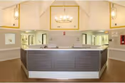 Interior view of a senior living facility reception area with a central desk featuring two sinks. The space has high vaulted ceilings with hanging light fixtures, beige walls, and a brown floor. There are numbered doors and windows on the walls behind the desk.