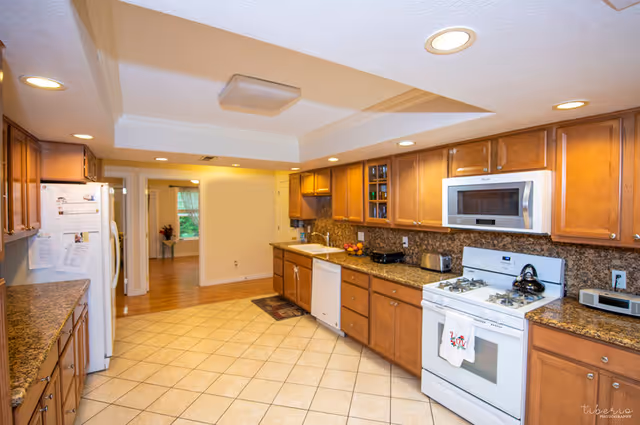 Spacious kitchen with wooden cabinets, granite countertops, a white gas stove and dishwasher, and tiled floor under recessed lighting.