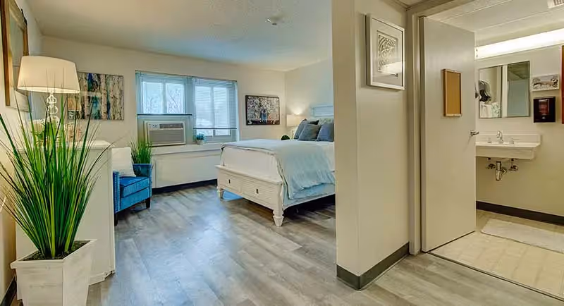 A bright senior living bedroom with a large bed dressed in light blue bedding, a blue armchair, a tall floor lamp, and a large potted plant. The room has wood flooring and a window with blinds. To the right, there is a partial view of a bathroom with a sink, mirror, and tiled floor.