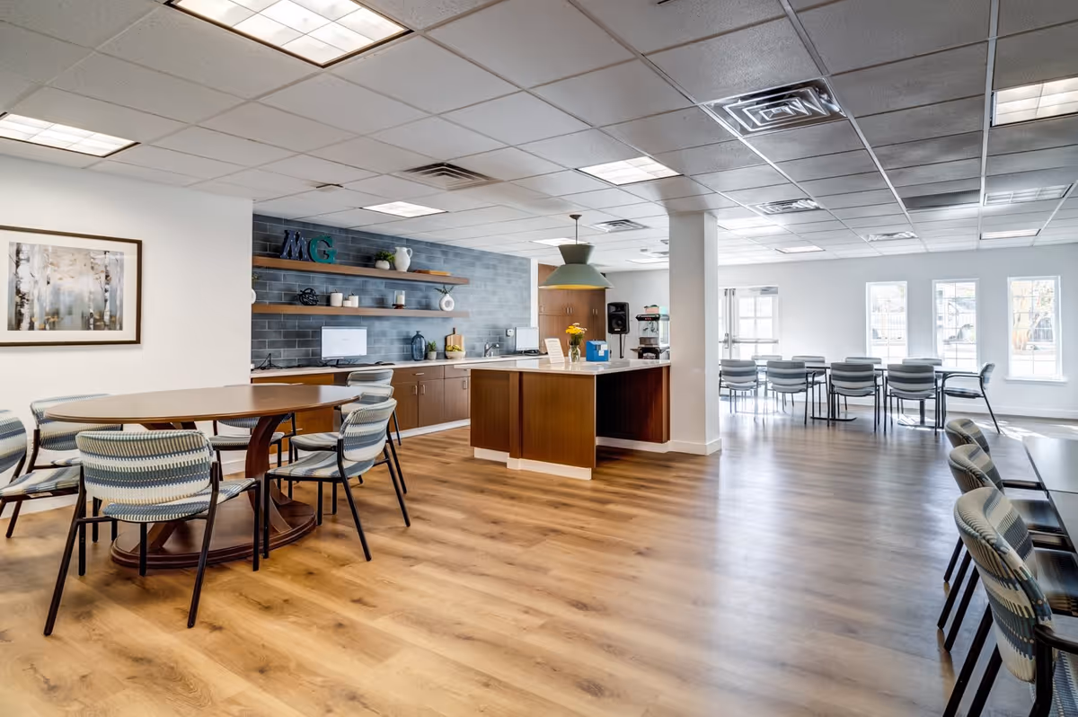 A spacious, well-lit common area with wooden flooring featuring multiple seating arrangements including round and rectangular tables with chairs. The room includes a kitchen area with wooden cabinets, open shelves with decorative items, and a central island. Large windows allow natural light to fill the space.