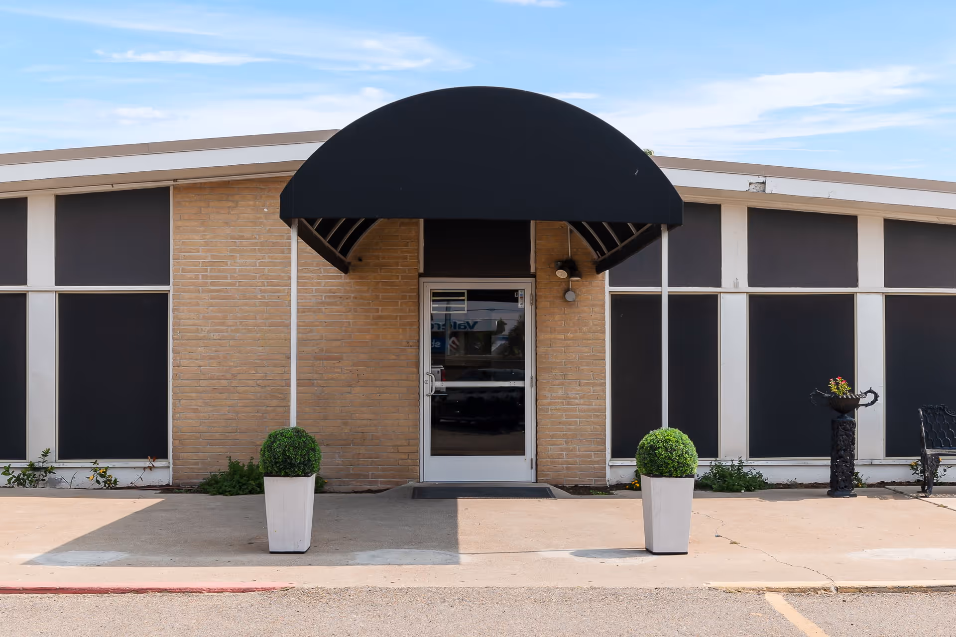 Entrance of a building with a black awning above a glass door, flanked by two white planters with green shrubs. The building has beige brick walls and large black window panels. There is a small black decorative planter and a bench to the right of the entrance.