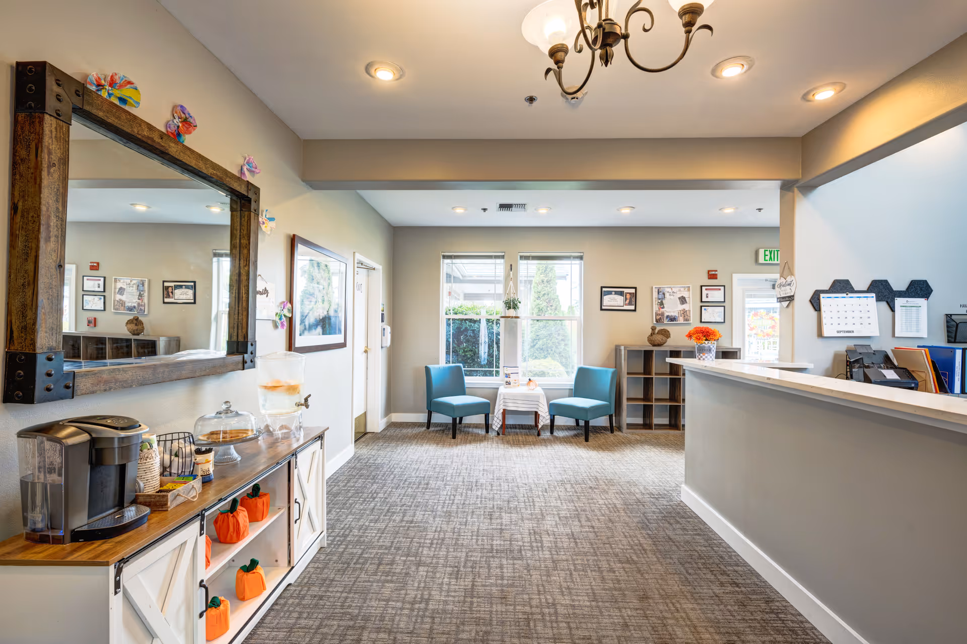 Bright reception lobby with a front desk, two blue chairs by a window, and a refreshment station with a mirror on the wall.