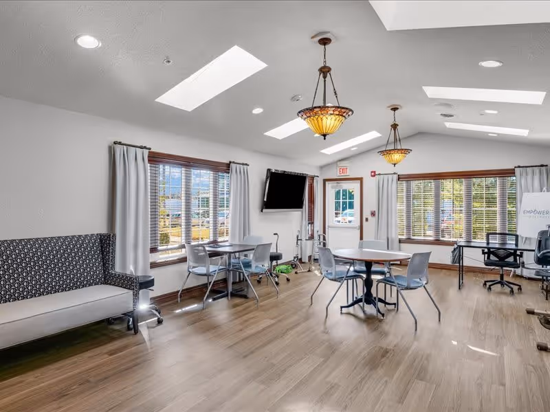 A bright and spacious common area with wooden flooring, multiple round tables with chairs, a patterned bench, large windows with white curtains, two hanging light fixtures, a wall-mounted TV, and an exit door. The room has skylights and recessed lighting in the ceiling.