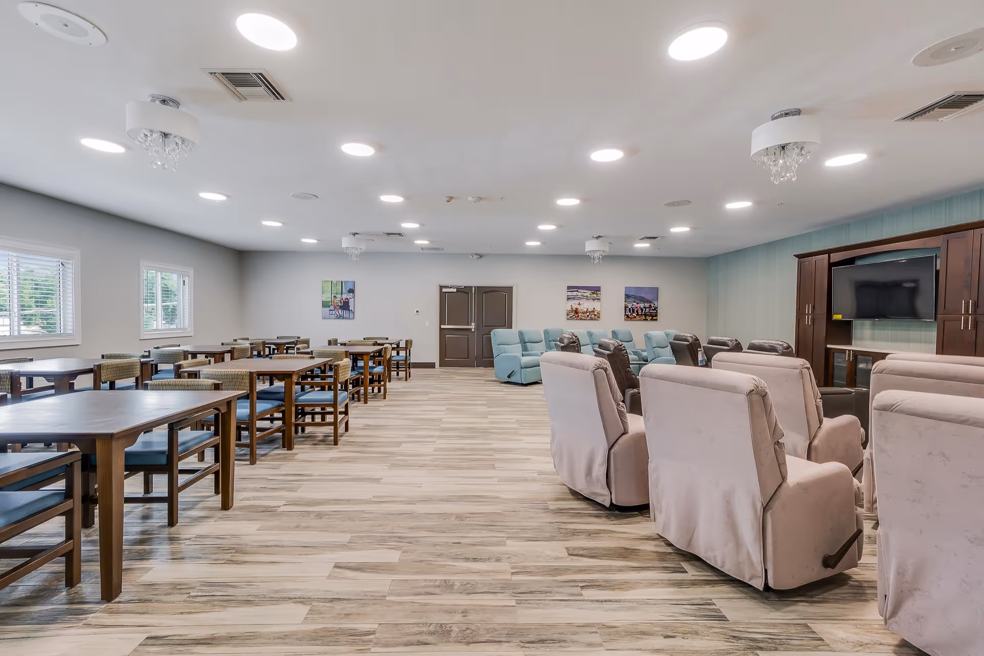 Large bright common room with rows of dining tables on the left and several recliner chairs facing a TV cabinet on the right.