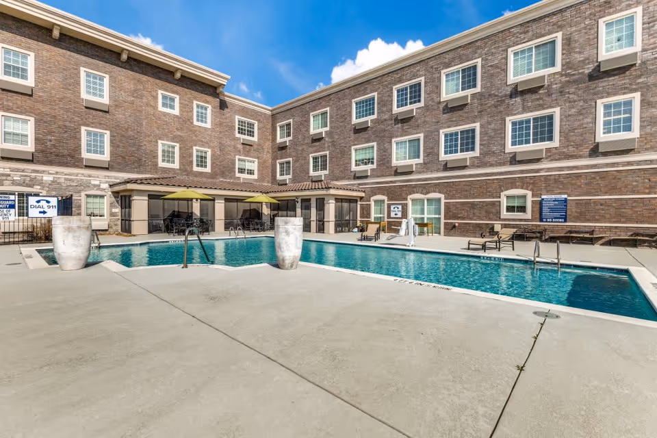 Outdoor swimming pool area at The Pearl Senior Living at Lewisville with lounge chairs, tables with umbrellas, and a three-story brick building in the background under a blue sky with some clouds.