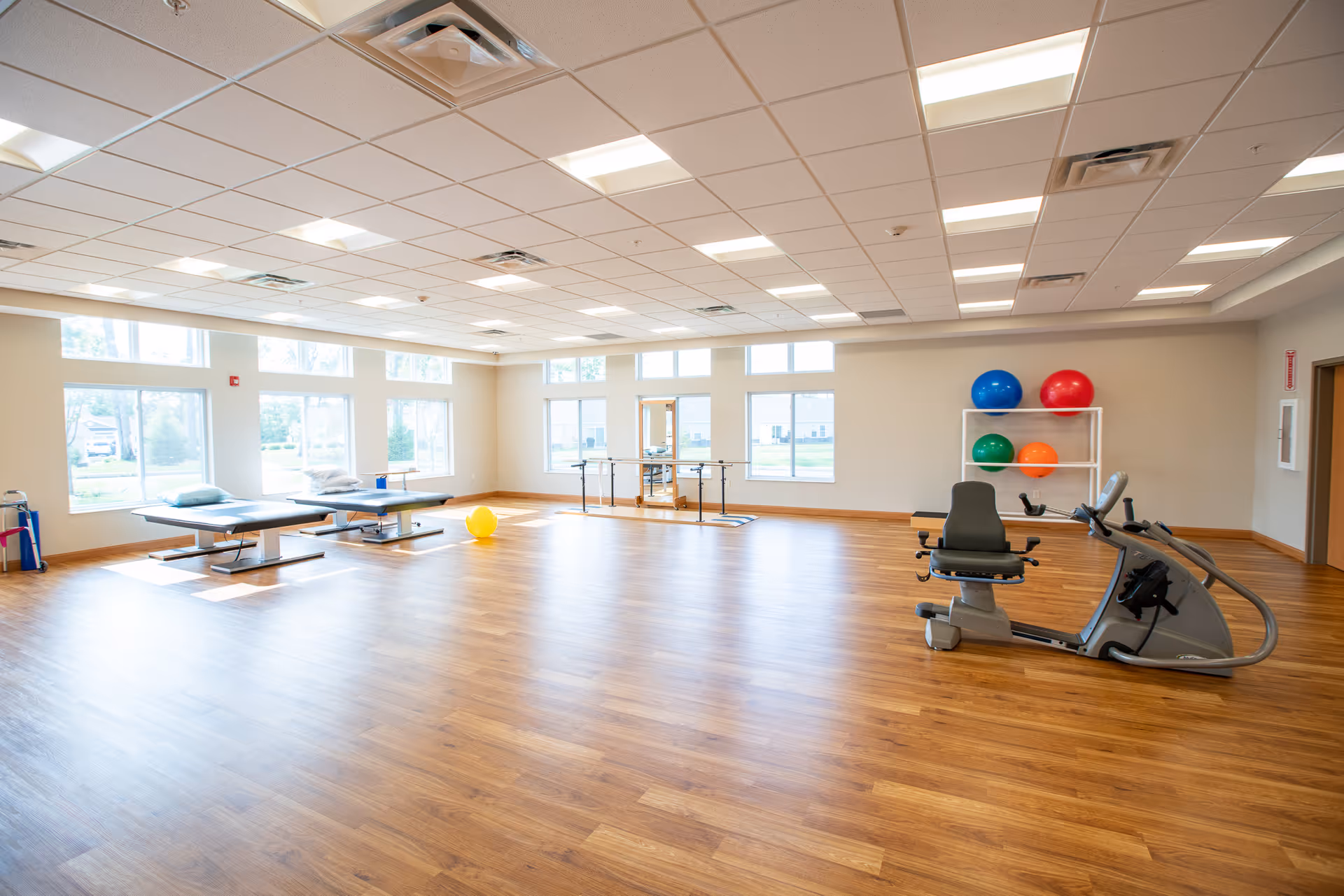 A spacious rehabilitation room with wooden flooring, large windows letting in natural light, two therapy tables with pillows, a stationary exercise bike, parallel bars for walking exercises, and a rack holding four colorful exercise balls.