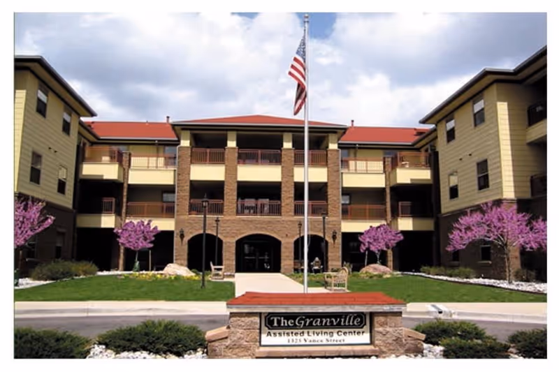 Front exterior view of The Granville Assisted Living Center building with a three-story structure, a central entrance, balconies, an American flag on a flagpole, and landscaped grounds with green grass and purple flowering trees.