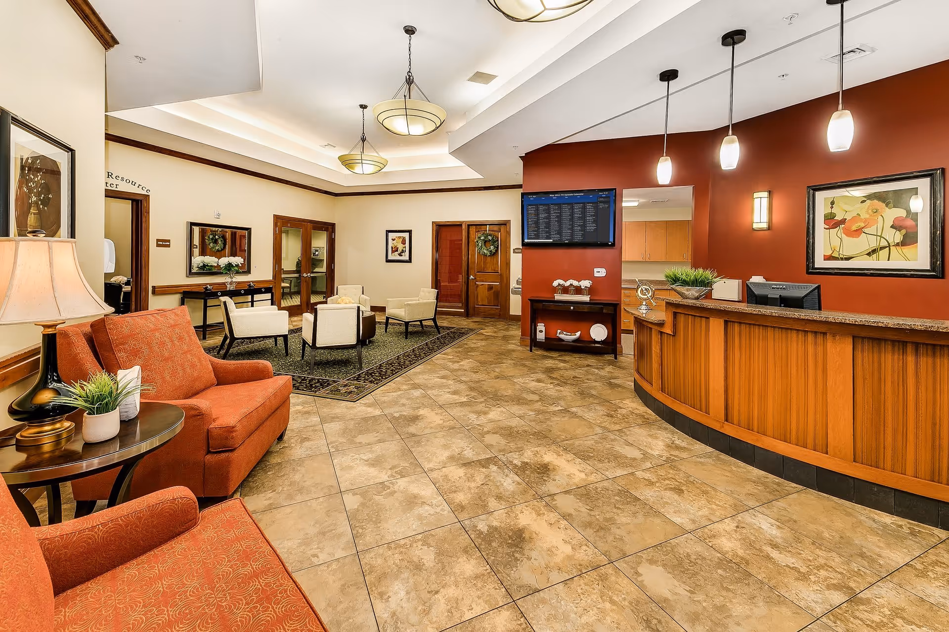A spacious and well-lit reception area with a wooden front desk on the right side, pendant lights hanging above it, and a wall-mounted TV screen. The room features beige tiled flooring, a seating area with orange upholstered chairs and a side table with a lamp and plant on the left, and additional seating with cream-colored chairs and a dark rug in the background. The walls are painted in warm tones with framed artwork and a door decorated with a wreath.