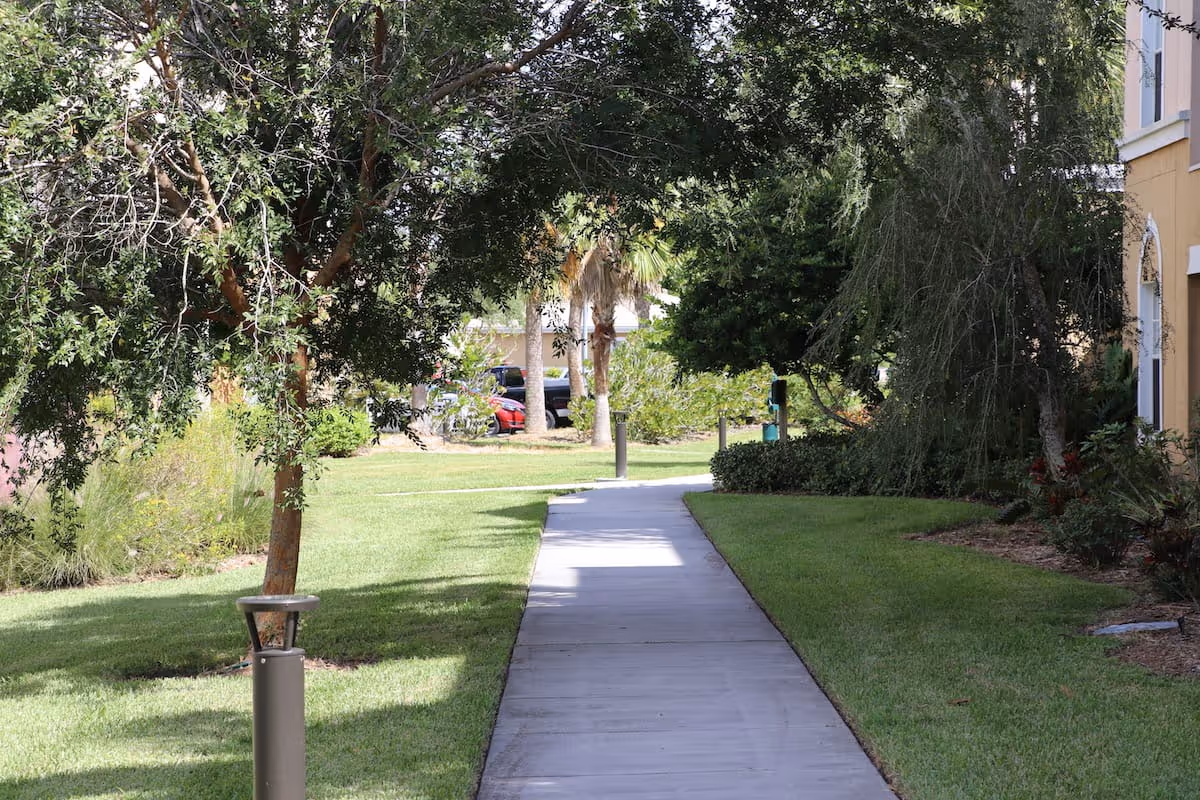 A paved walkway surrounded by green grass and trees leading through a landscaped outdoor area next to a yellow building.