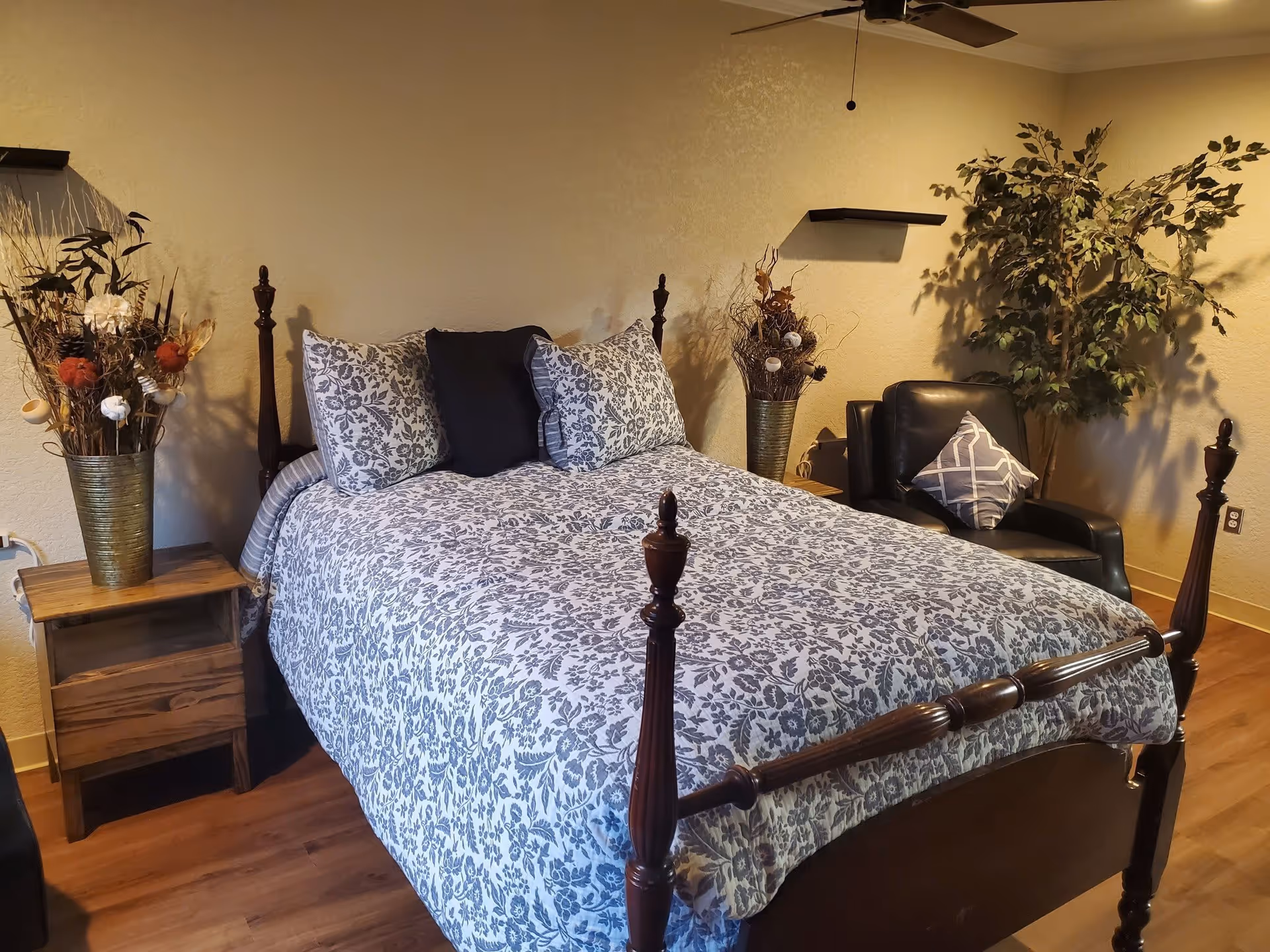 A cozy bedroom with a wooden four-poster bed covered in a floral patterned bedspread and pillows. There is a wooden nightstand with a decorative vase of dried flowers on the left side of the bed. On the right side, there is a black leather armchair with a patterned cushion and a large artificial plant behind it. The walls are painted beige and there is a ceiling fan above.