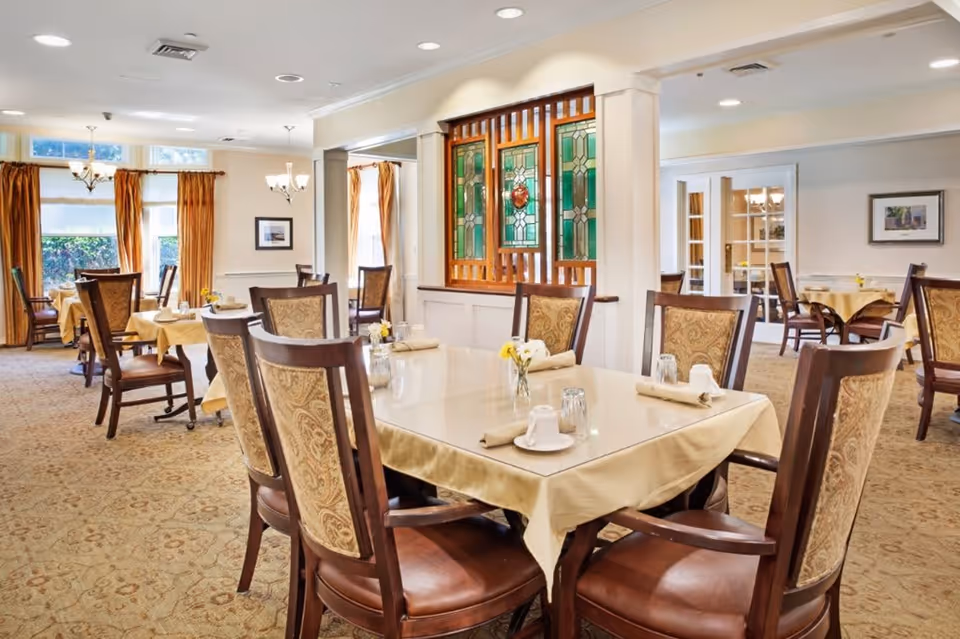 A well-lit dining room with multiple tables covered in beige tablecloths, each set with napkins, cups, and small flower vases. The room features upholstered wooden chairs, large windows with orange curtains, and decorative stained glass panels on one wall.