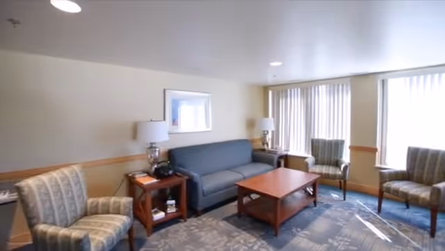 A cozy living room with a blue sofa, two patterned armchairs, a wooden coffee table, two side tables with lamps, and large windows with vertical blinds letting in natural light.