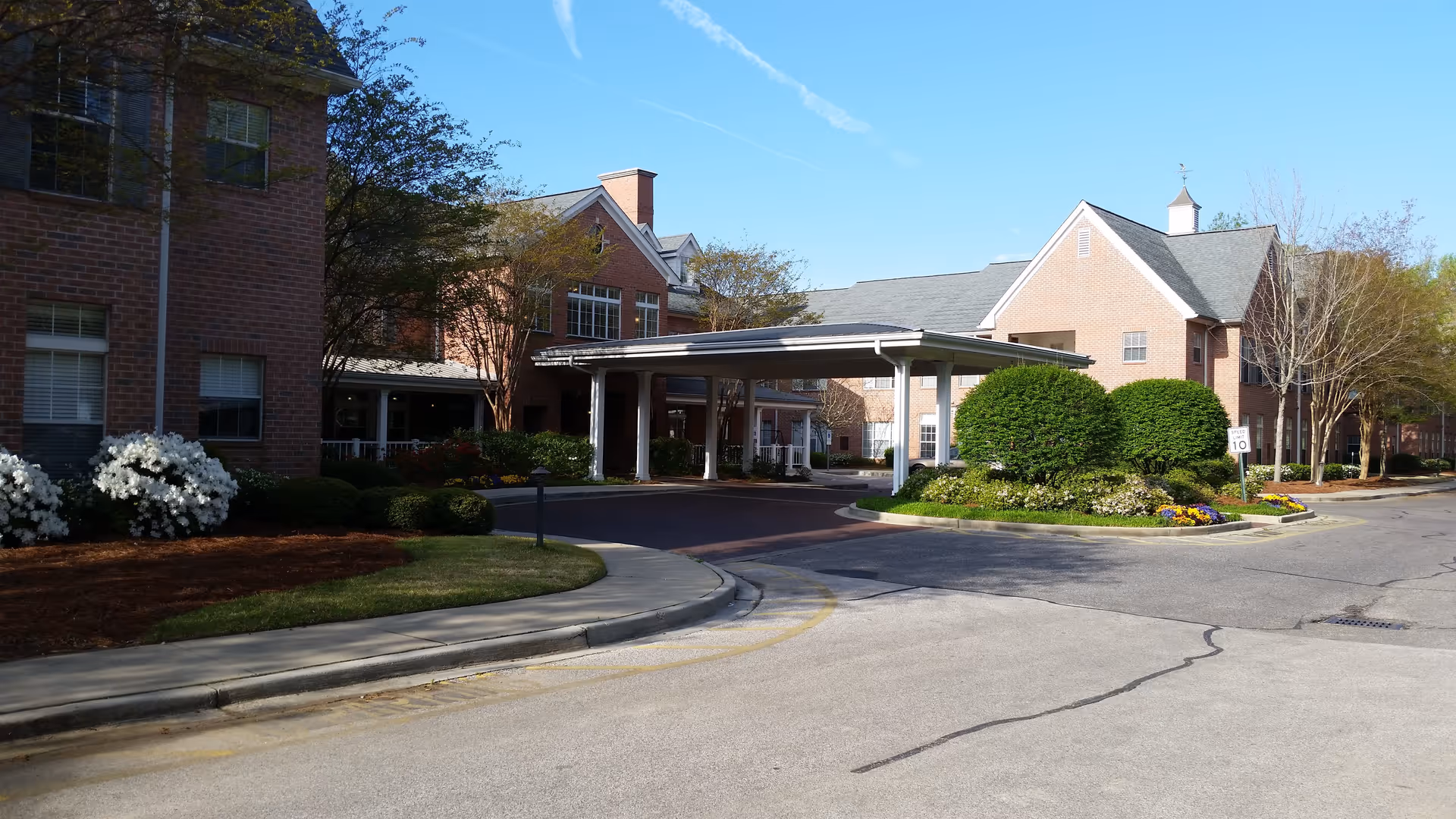 Exterior view of a senior living facility with brick buildings, a covered entrance, landscaped bushes, flowers, and a clear blue sky.