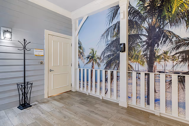 An indoor area with light gray wooden walls, a white door, a black coat rack, and a large wall mural depicting a tropical beach scene with palm trees and a sandy shore. The floor is covered with light wood-patterned tiles, and there is a white railing in front of the mural.