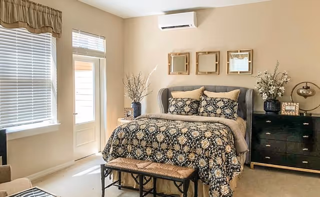A cozy bedroom with a large bed covered in a patterned black and beige comforter and matching pillows. The bed has a cushioned headboard and a bench at the foot. There are three square mirrors hanging above the bed. To the right, there is a black dresser with decorative items including a lamp and vases with flowers. A window with blinds and a door letting in natural light are on the left side of the room. The walls are painted beige and there is an air conditioning unit mounted above the bed.