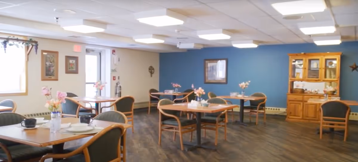 A dining room with several wooden tables and chairs arranged neatly. Each table has a small vase with pink flowers and some have cups and plates. The room has a blue accent wall with a framed picture and a wooden cabinet displaying dishes. The floor is dark wood, and there is a door and window on the left side letting in natural light.