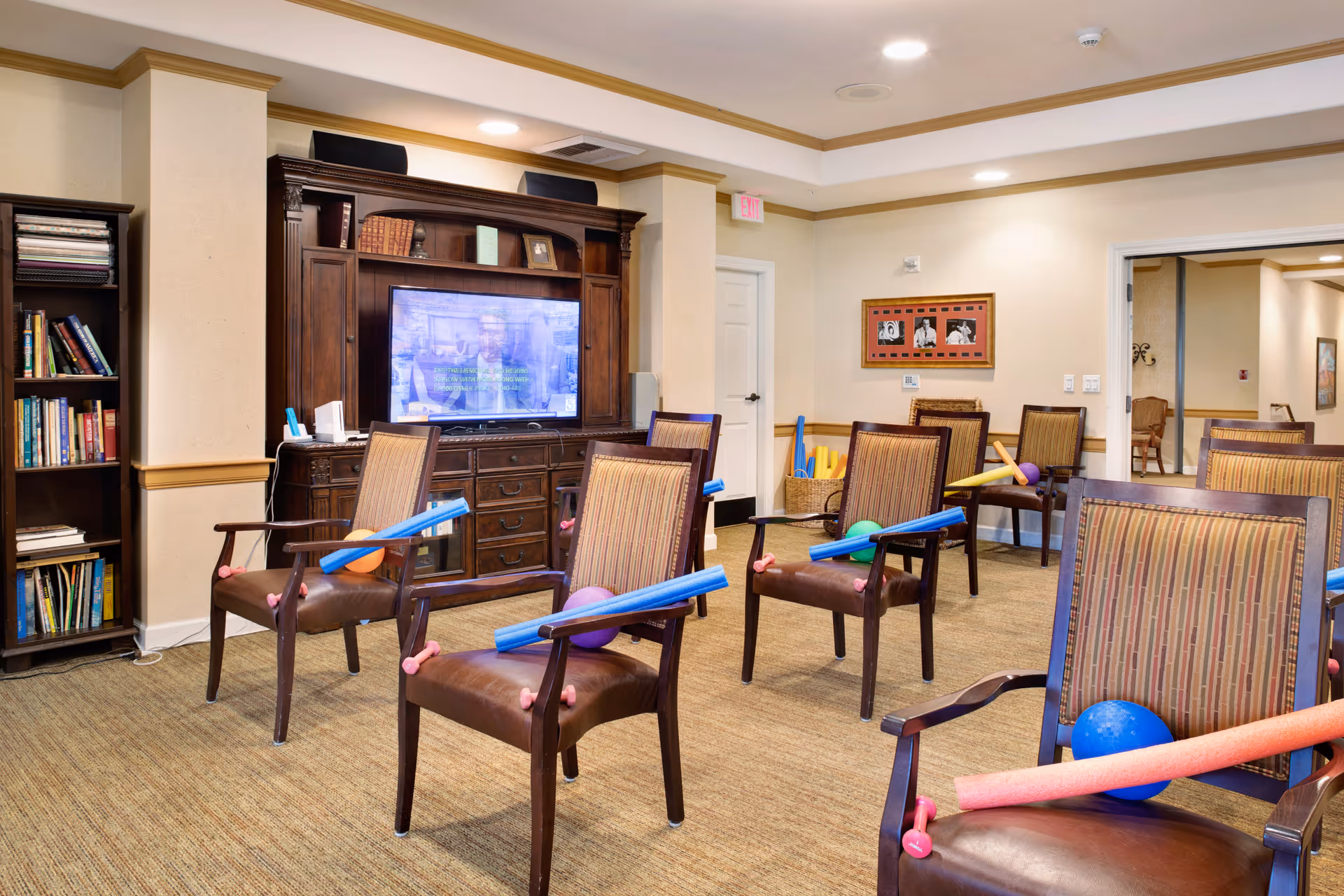 Well-lit communal activity room with rows of chairs facing a large TV and exercise equipment like pool noodles and exercise balls.