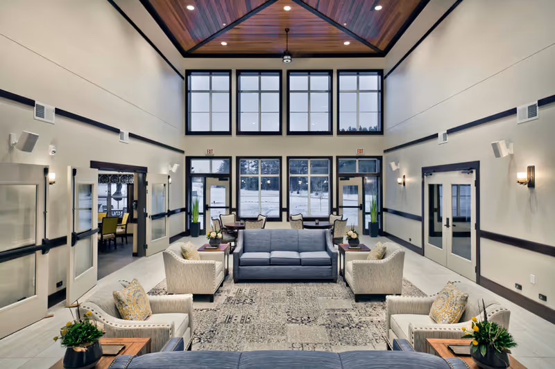 Bright, high‑ceilinged communal living room with sofas, armchairs, side tables and large windows overlooking the outside.