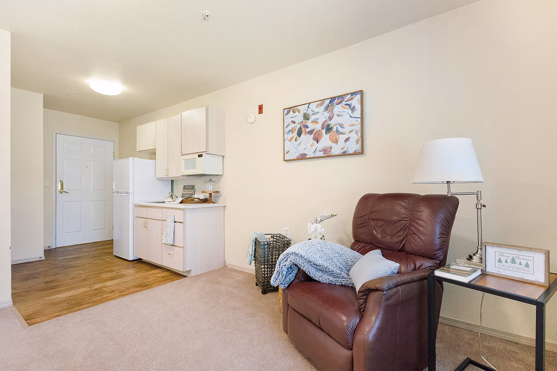 A small living area with a brown leather armchair draped with a blue blanket and a pillow. Next to the chair is a side table with a lamp, books, and a decorative sign. The background shows a compact kitchen with light wood cabinets, a white refrigerator, microwave, and countertop with some items. The floor transitions from carpet in the living area to wood in the kitchen. A framed artwork with leaves hangs on the wall above the chair.